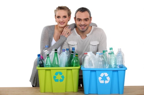 In this image, a smiling woman and man are positioned behind two plastic recycling bins placed on a wooden surface against a plain white background. The woman has light skin, blonde hair tied back, and is wearing a light grey cardigan, with her left hand resting on the man's right shoulder. The man has short dark hair, light skin, and is wearing a white long-sleeved shirt with the sleeves rolled up. The bin on the left is lime green with a recycling symbol on the front, filled with assorted transparent plastic bottles, including some with coloured caps. The bin on the right is blue, also featuring a recycling sign, and contains similar clear plastic bottles. The scene suggests a focus on recycling practices related to waste management, which aligns with services provided by Commercial Waste Removal Kilburn in the local area, possibly serving businesses or residents in the Kilburn postcode region. The lighting is bright and even, clearly highlighting the details of the bottles and the recycling bins, emphasizing environmentally conscious waste segregation.