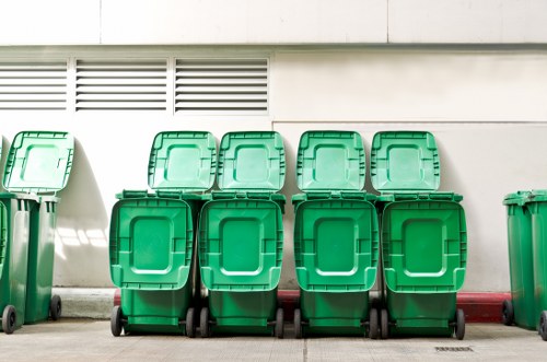 A row of six green wheelie bins with hinged lids, made of durable plastic with smooth surfaces, are positioned on a pavement in front of a white wall featuring ventilation panels. The bins are arranged side by side, and four of them are placed directly in front of a red line painted on the pavement. The environment appears to be an outdoor urban setting, possibly near a commercial or residential building in Kilburn, with clear lighting indicating daytime. The image emphasizes waste management and rubbish removal, aligning with services offered by Commercial Waste Removal Kilburn. The bins’ general condition appears clean and well-maintained, and the scene captures a typical rubbish collection area, ready for servicing or disposal, reinforcing the focus on efficient waste management for local businesses or residents.