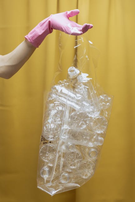 A person wearing a pink rubber glove holds a transparent plastic bag filled with clear crushed plastic bottles and containers. The bag is suspended against a yellow background, emphasizing the recycli